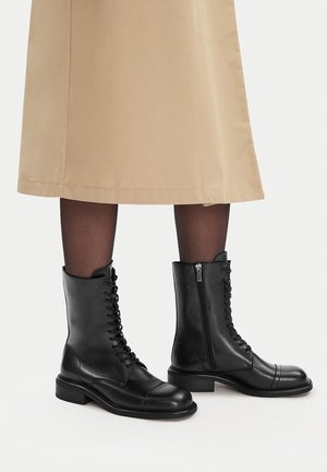 Czarne skórzane botki na sznurowadła z bocznym suwakiem noszone z przezroczystymi czarnymi rajstopami i beżową spódnicą.