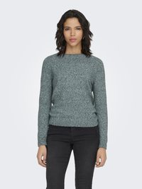 Femeie cu păr ondulat până la umeri, purtând un pulover verde texturat și pantaloni negri, stând pe un fundal simplu și deschis la culoare.