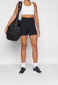 Black athletic shorts and a white sports bra, paired with black sneakers and ankle socks. The model holds a black gym bag with a mesh design.