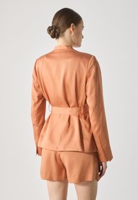 Peach-colored blazer with a peplum design, long sleeves, and a belted waist, paired with matching high-waisted shorts. Smooth texture.