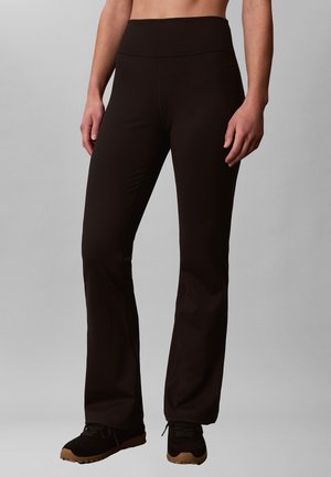 Mujer con pantalones negros acampanados de cintura alta y zapatillas negras, de pie frente a un fondo gris liso.