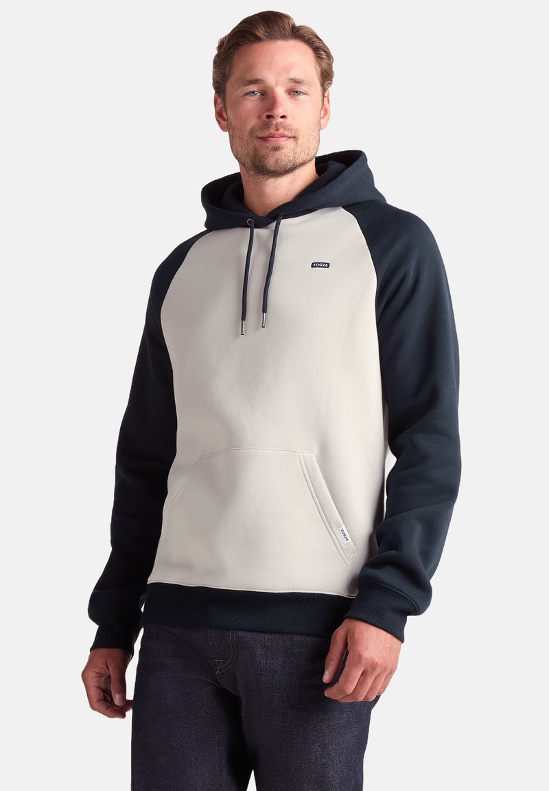 Hooded sweatshirt in cream with navy sleeves, featuring a front pocket, drawstrings, and a small logo on the chest. Soft, textured fabric.