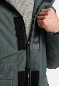 Grön ytterjacka med dragkedja framtill, kardborrefästen och en gråtexturerad tröja under. Har slät tyg och svarta detaljer i hårdvara.