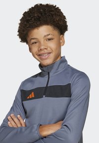 Cima deportiva de color gris con cremallera en el cuello, con una franja horizontal negra y un logo naranja en el lado izquierdo del pecho. Textura suave, mangas largas.