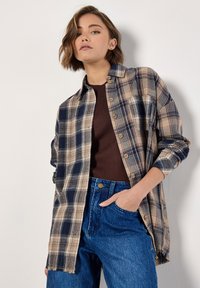 Flannel shirt in navy, beige, and light brown plaid pattern with wooden buttons, long sleeves, and frayed hem, worn over a brown top.