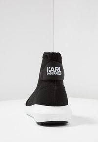 Black high-top sneaker with a knit upper, white sole, and a rear logo tag featuring "Karl Lagerfeld." Textured fabric with a smooth heel.