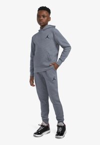 Conjunto de sudadera con capucha gris y pantalones de chándal, con un logo Jumpman negro. Tejido suave con un corte relajado. Zapatillas en negro con detalles en rojo.