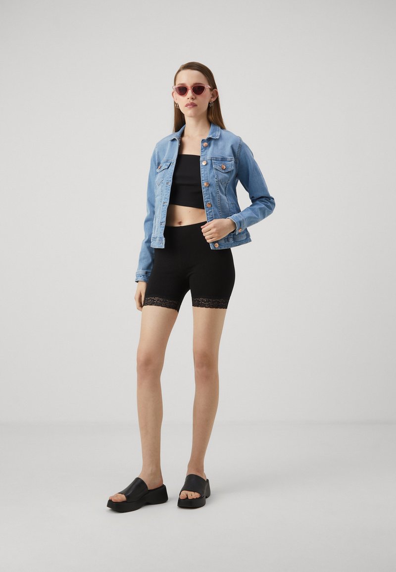 Lichtblauwe denimjas met koperen knopen, zwart crop top, zwarte shorts met kantafwerking, en zwarte gesloten slipper-sandalen.