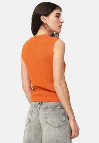 Top en tricot sans manches orange avec une finition texturée, présentant un design ajusté et un ourlet côtelé, associé à un jean taille haute gris décoloré.