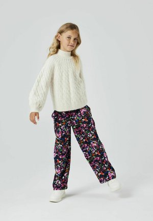 Pull en tricot blanc avec col montant, associé à un pantalon noir à motif floral aux couleurs rose, violet et vert. Baskets blanches.