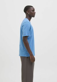 Tricou din bumbac albastru, cu mâneci scurte, croială relaxată, textură fină, gât rotund. Purtați cu pantaloni gri. Vedere din profil.