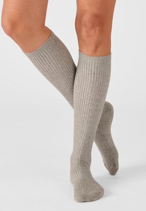Due gambe incrociate che indossano calze alte fino al ginocchio a coste grigio chiaro su uno sfondo chiaro uniforme.
