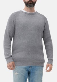 Mann, der einen gerippten grauen Strickpullover über einem weißen Unterhemd und hellblauen, zerrissenen Jeans trägt, vor einem einfarbigen Hintergrund.