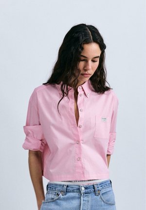 Chemise rose à manches courtes en coton, dotée d'une poche avant et de manches retroussées. Associée à un jean en denim bleu clair.