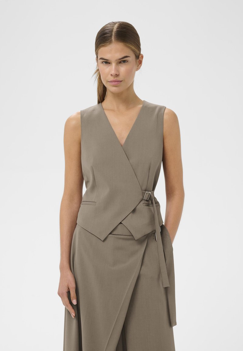 Bež brezrokavnik z V-izrezom, oprijetim krojem, stranskimi žepi in pasom s srebrno sponko, narejen iz gladkega blaga.