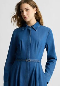 Denim kleit kaeluse ja vöökohtadega, mis on kaunistatud hõbedase pandlaga, pikkade varrukatega ja flared seelikuosaga keskmiselt sinises värvitoonis.