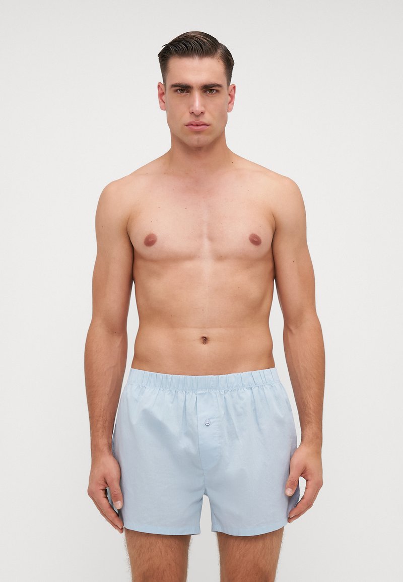 Boxer shorts in cotone azzurro chiaro con vita elasticizzata e chiusura anteriore con bottone, esposti su un modello maschile senza camicia contro uno sfondo neutro.