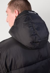 Zwarte pufferjack met een gestructureerde capuchon. Het materiaal heeft een glanzende afwerking, met doorgestikte naden en een merklabel bij de hals.