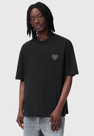 Camiseta negra de algodón con un pequeño logo de corazón en el lado izquierdo del pecho, mangas cortas y un corte relajado. El detalle texturizado añade profundidad.