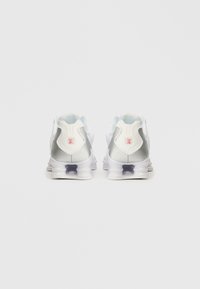 Ténis atléticos brancos e prateados com colarinho acolchoado, parte superior em malha, sola transparente e detalhe em vermelho no calcanhar.