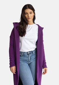 Fioletowy kardigan z kapturem wykonany z miękkiej tkaniny, z długimi rękawami i otwartym przodem. Noszony na białej koszulce i niebieskich dżinsach.