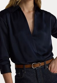 Marineblauwe satijnen blouse met een diepe V-hals, lange mouwen opgerold bij de manchetten, gecombineerd met een bruin leren riem en donkere jeans.