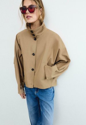 Žena v nadrozmernej béžovej bundě na gombíky s vysokým golierom, veľkými vreckami, modrými džínsami a nadrozmernými štvorcovými červenými slnečnými okuliarmi.