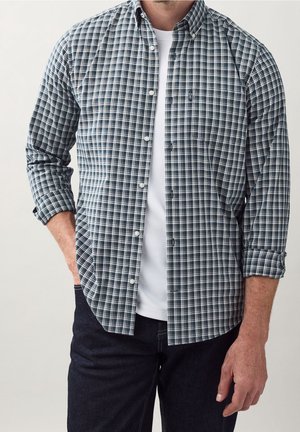 Mężczyzna w niebiesko-białej koszuli w kratę z długim rękawem, na białym t-shircie i w ciemnych dżinsach, z jedną ręką w kieszeni.