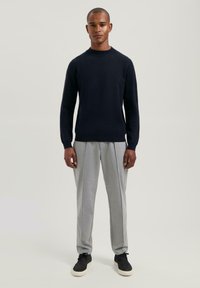 Hombre de pie vistiendo un suéter negro de manga larga, pantalones grises claros con pliegues y zapatillas negras con suelas blancas sobre un fondo plain.