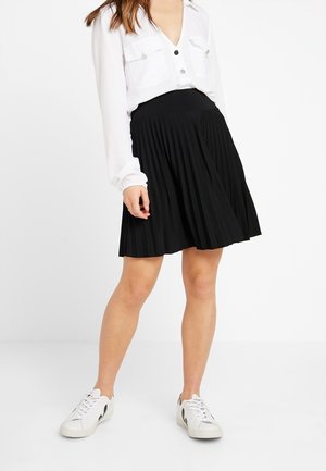 Jupe plissée noire avec une taille cintrée, portée avec une chemise blanche à boutons et des baskets blanches, mettant en avant la texture et le mouvement.