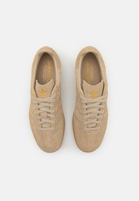 Béžové semišové tenisky se světle šedými tkaničkami, s třemi nenápadnými pruhy a zlatým logem Adidas na jazyku a vložce.