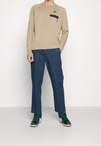 Osoba w beżowej bluzie Nike z kieszonką na zamek na piersi, w niebieskich szerokich dżinsach i zielono-czarnych białych sneakersach, stoi przy białej ścianie.