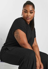 Czarna, strukturalna stylizacja z krótkimi rękawami i luźnym krojem; widoczny tatuaż na ramieniu. Prosty design, bez dodatkowych wzorów czy akcentów.