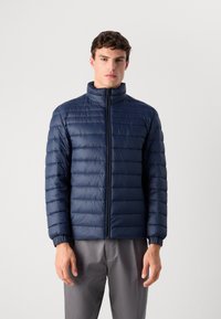 Young man wearing a navy blue quilted jacket with a front zipper and gray pants, standing against a plain light background.
