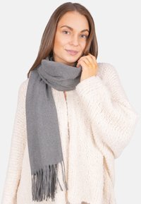Femme portant un gros pull crème et une longue écharpe grise enroulée autour de son cou, avec une main touchant l'écharpe.