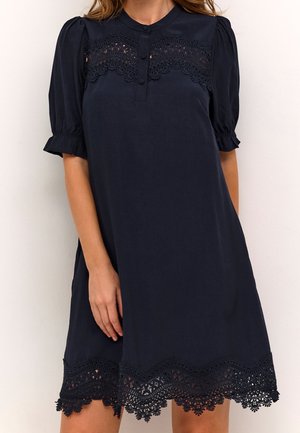 Femme portant une robe bleu marine avec des détails en dentelle sur la poitrine et l’ourlet, des manches courtes bouffantes et une patte de boutonnage à l’avant.