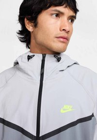 Chaqueta Nike de color gris claro con detalles en negro y cierre con cremallera. Cuenta con capucha y logo en el pecho. Textura suave, diseño atlético.