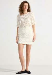 Vit klänning med texturerad blommönster, korta ärmar och kroppsnära passform. Genomskinligt ytterlager över en solid bodice. Svarta stickade skor.