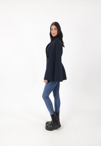 Blazer capea azul marino con un collar alto, combinado con jeans skinny azules y botas de plataforma negras; presenta un tejido fluido y un diseño estructurado.
