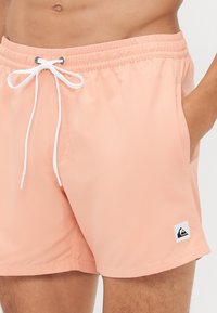 Shorts de bain pêche clair en tissu lisse, dotés d'une taille élastique avec cordons blancs et de poches latérales. Patch logo en bas.