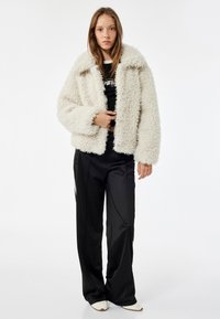 Faux fur jakke i off-white, cropped design med en bred krave og lange ærmer, parret med sorte, løstsiddende bukser. Glat, skinnende tekstur.