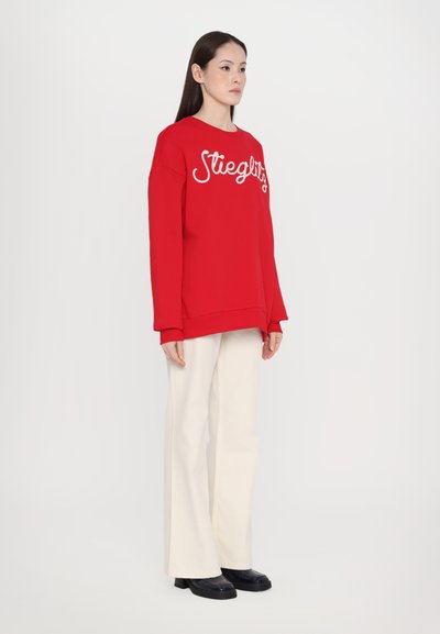 Sudadera roja con texto en blanco, combinada con pantalones de pierna ancha beige claros y zapatos de plataforma negros. Tejido suave, corte relajado.