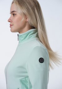Mintgrön zip-up-jacka med hög krage. Har en logotyp på vänster ärm och en slät textur. Sidoficka synlig.
