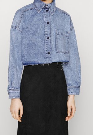 Kvinde iført en kort blå denimjakke med sorte knapper og en stor brystlomme, kombineret med en sort nederdel med høj talje.