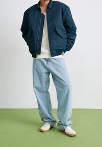 Giubbotto bomber navy con tasche frontali, abbinato a jeans baggy azzurri e sneakers bianche con dettagli color sabbia, su un pavimento verde.