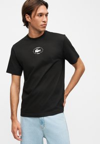 Άνδρας που φοράει μαύρο T-shirt Lacoste με λευκό λογότυπο και ανοιχτόχρωμο μπλε τζιν, στέκεται με το ένα χέρι πίσω από την πλάτη, κοιτάζοντας αριστερά.