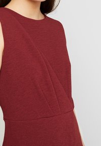 Burgundowa sukienka bez rękawów z teksturowanej tkaniny z ukośnymi szwami. Okrągły dekolt i dopasowany krój.