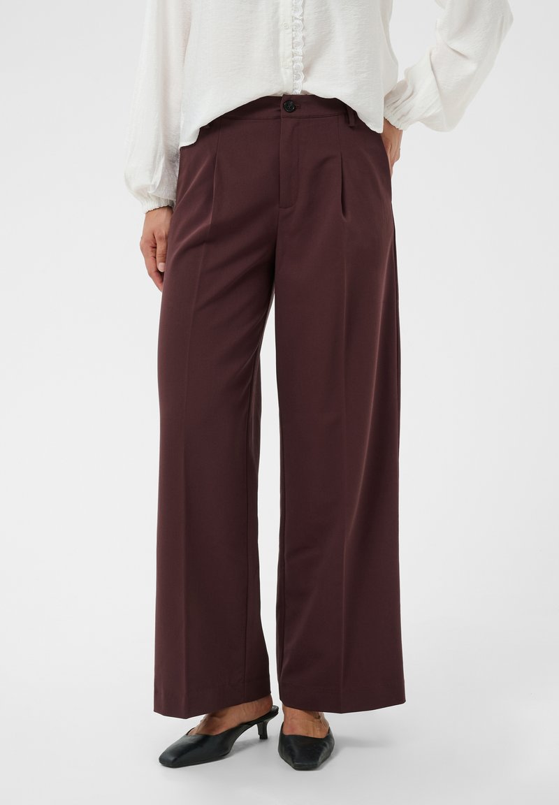 Femme portant un pantalon marron foncé taille haute, jambes larges avec plis devant, et un chemisier blanc, associé à des chaussures noires à talons bas.