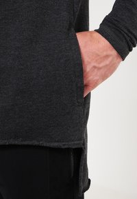 Rechte Hand teilweise in der Seitentasche eines dunkelgrauen Langarmshirts über einer schwarzen Hose mit Reißverschlusstasche.