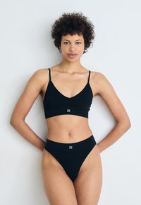Mustat ribbed bralette ja yhteensopivat thongit, joissa on leveä elastinen nauha "HUGO"-logolla. Säädettävät olkaimet, sileä rakenne.